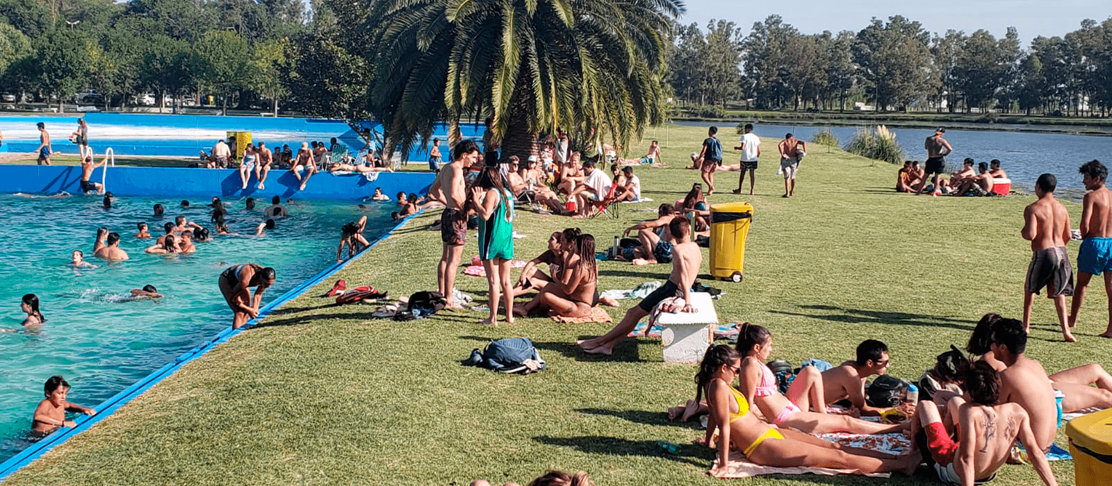 Gente sentada sobre el cesped a orillas de la pileta